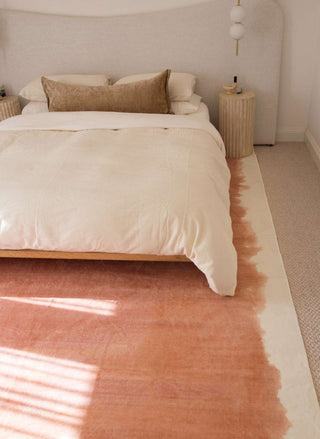 Neatly made bed with beige bedding in a bedroom with a pinkish-orange rug.